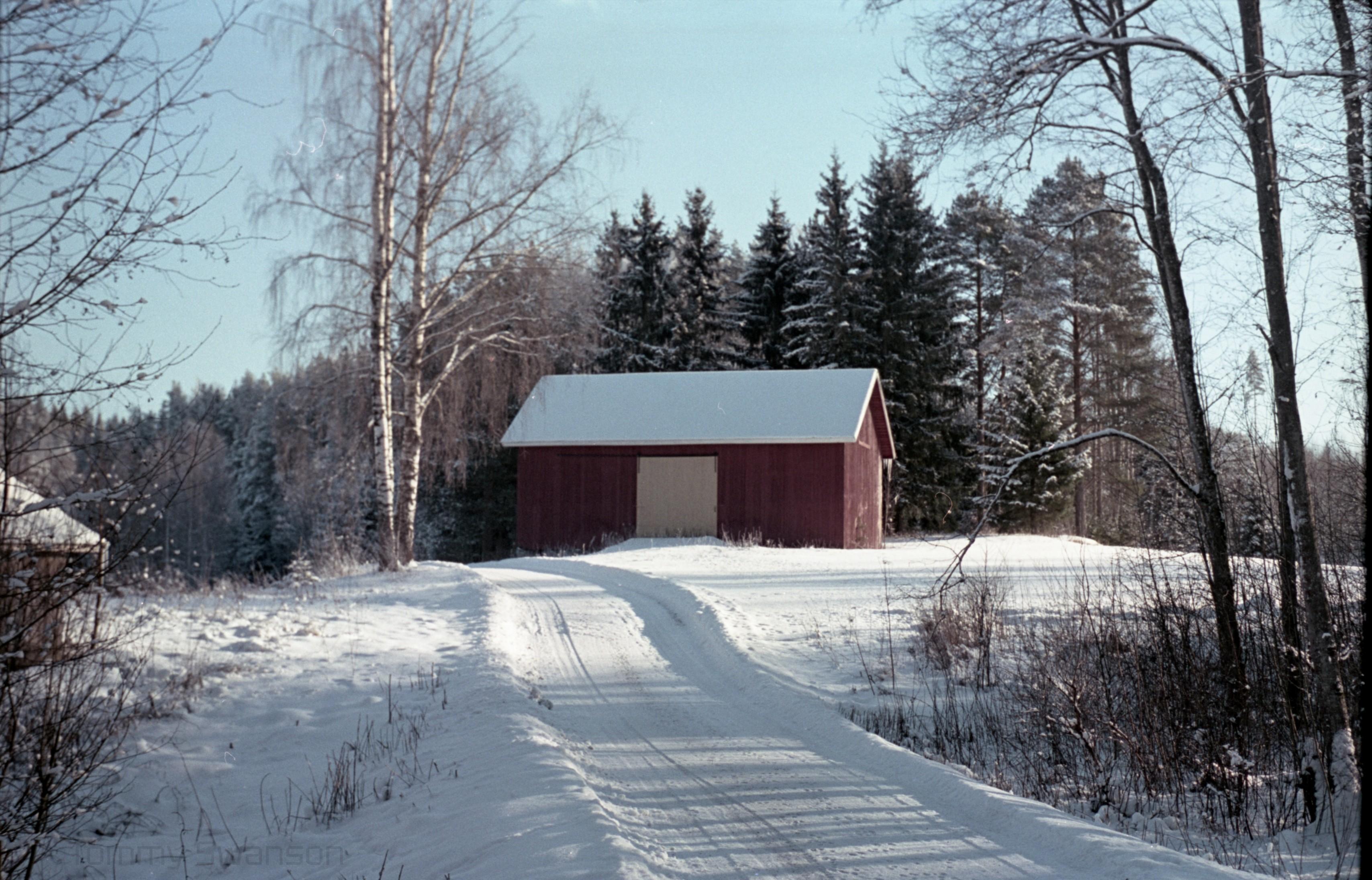 A wooden red barn in front of a grove on top of a hill. A plowed road leads past the barn.