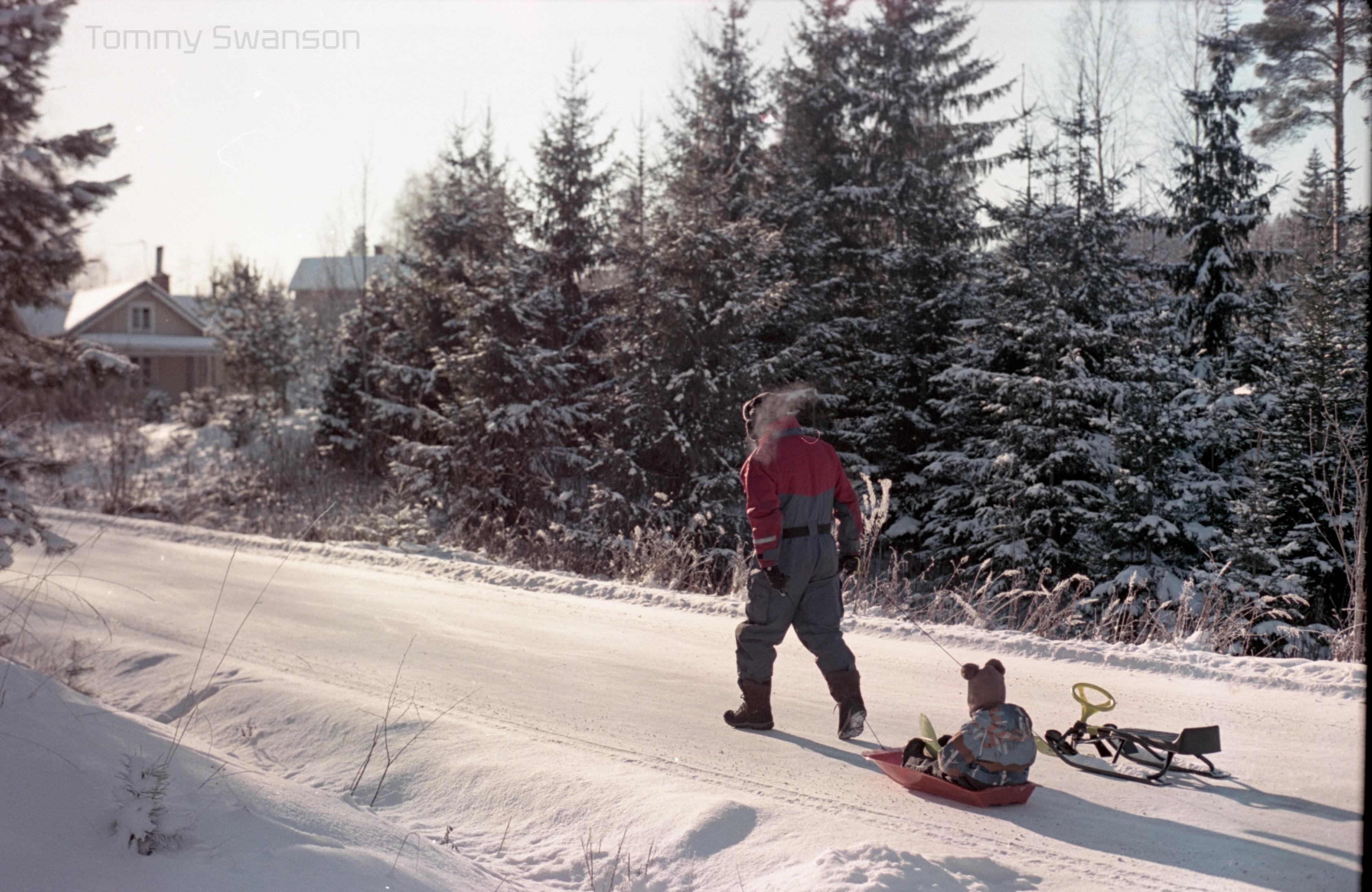 A man dressed in overalls pulls a preschool-aged child in a sled with one hand and an empty sled with his right hand on a plowed road.