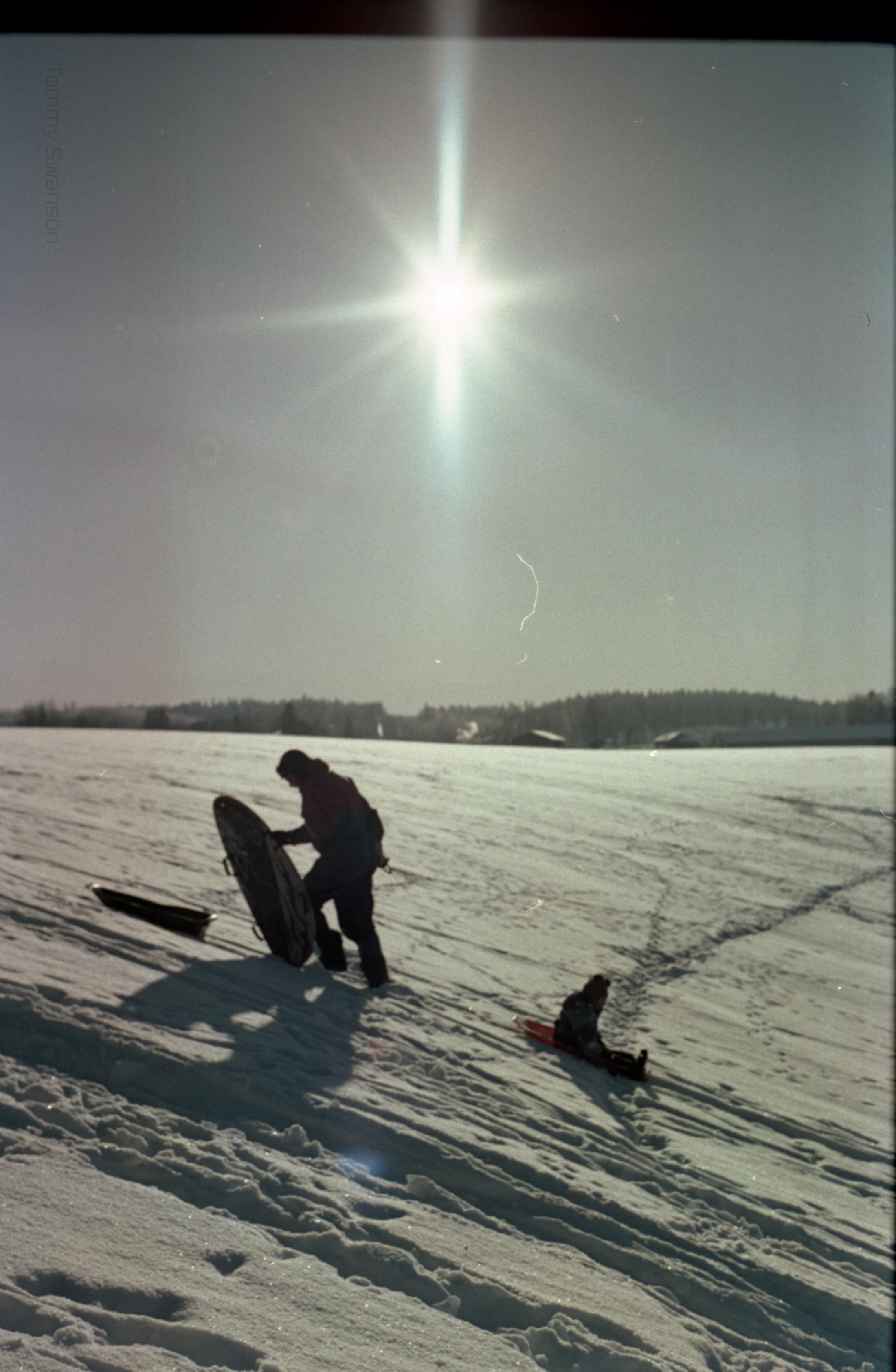 A man wearing overalls pulls a preschool-aged child up a hill on a sled. Directly behind them is the sun in the sky.