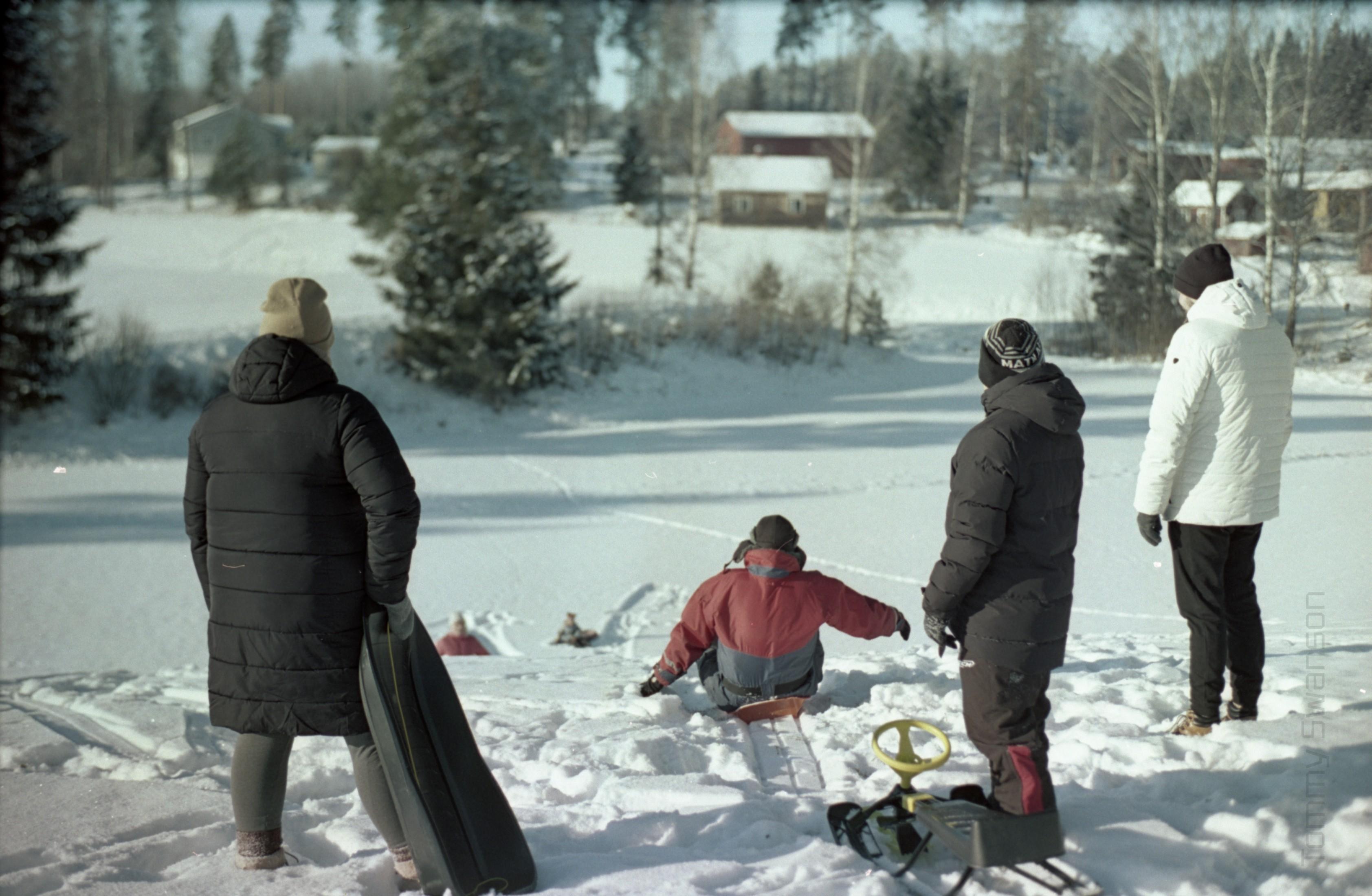 A man wearing overalls is sledding down a hill. There are two people at the bottom of the hill and three people watching at the top.